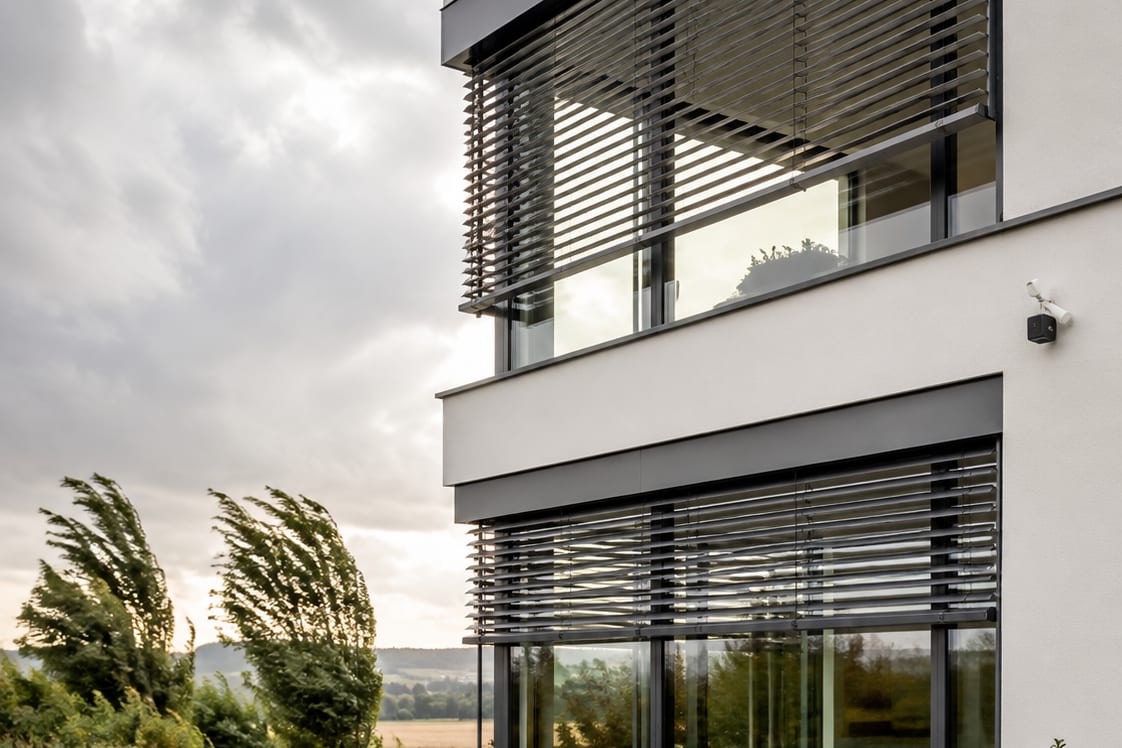 Hausfassade mit Außenjalousien aus dunklen Aluminiumlamellen, im Hintergrund vom Wind gebeugte Bäume